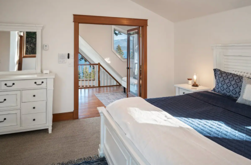 Bright bedroom with a white bed, dresser, and a view of a staircase leading to another room. Natural light fills the space.